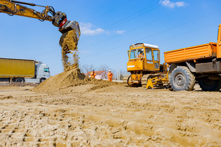 View on excavator and truck with mounted plate vibration compactor, as they are compacting, leveling sand for road foundation at building site, mechanization.の写真素材
