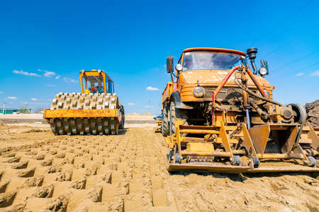 Road roller with spikes and truck with mounted plate vibration compactor are compacting, leveling sand for road foundation at building site.の写真素材