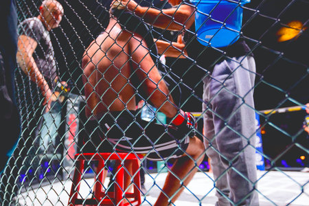 View through the wire of the fighting ring, trainer puts bag of ice on the fighter's neck to cool him down and wipes him with a towel.のeditorial素材