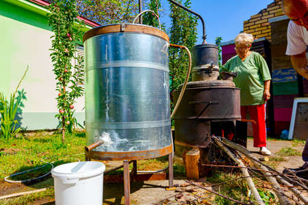 Woman is manually turning lever to mix fruit marc in boiler of homemade distillery made of copper, making brandy and man throws dry branches into the firebox of a homemade distillery for making alcoholic beverages.の写真素材