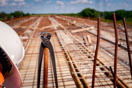 White safety helmet and metal pliers for binding wire reinforcement hung on a plastic tube, square rusty armature, placed for the strength of the bridge on the construction site.の写真素材