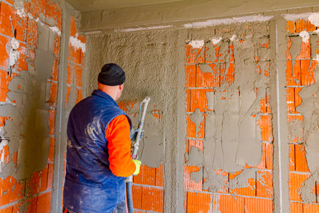 Worker man is holding mortar hose in his hand and plastering under pressure interior brick walls in...