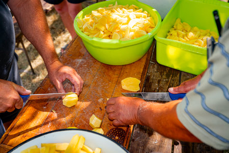 Teamwork of two farmer cookers cut potatoes using knives into slices on the wooden table prepare it...
