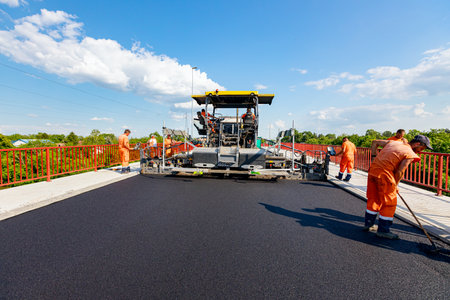 Zrenjanin, Vojvodina, Serbia - June 8, 2021: View on machine for laying asphalt, spreading layer of hot tarmac on prepared ground a few workers are using shovels to level it.のeditorial素材