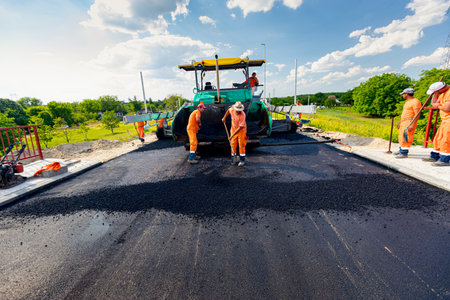 Zrenjanin, Vojvodina, Serbia - June 8, 2021: View on machine for laying asphalt, spreading layer of hot tarmac on prepared ground a few workers are using shovels to level it.のeditorial素材