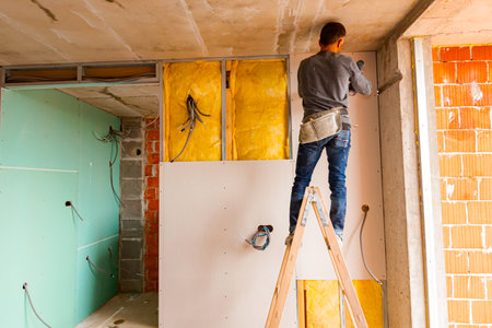 Worker on wooden ladder holds professional manual power tool ACU drill and tightens the screws on plasterboards installed on the partition dry wall insulated with mineral wool.の写真素材