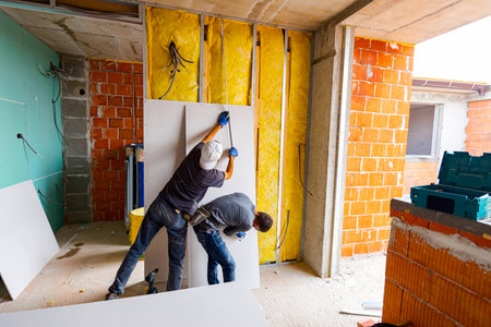 Teamwork, workers are measuring with a tape measure, checking wall dimensions before installing the plasterboards on the partition insulated wall. Building is under construction.の写真素材