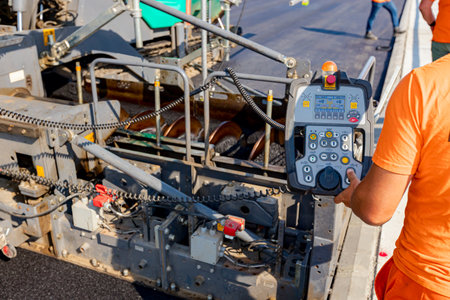 Operator, worker is holding industrial remote control, console with joystick to directing asphalt spreading machine, determines position for pouring, laying tarmac in right direction, road works on bridge under construction.の写真素材