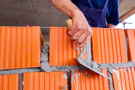 Worker is using spatula, trowel, to scrape off any excess mortar that spreads beyond the wall joint,...