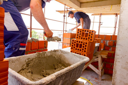 Mason, bricklayer worker grabs mortar from the plastic trough with spatula and smears red blocks to...