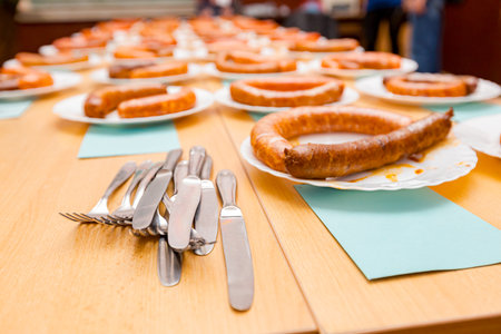 Bunch of cutlery, knifes and forks next to several white plates with two traditional sausages each, one cooked and other raw, exposed on exhibition table for estimation and review at food contest.の写真素材