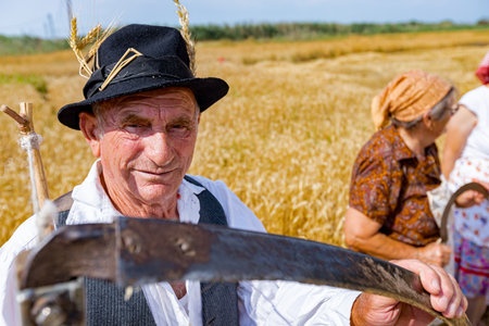 Muzlja, Vojvodina, Serbia, - July 02, 2022; XXXIX Traditionally mowing wheat. Portrait of senior farmer ready for harvest is holding scythe in front of field with mature grain.のeditorial素材