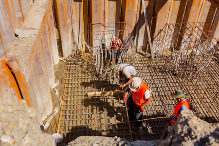 Shot above on workers surrounded by metal piles, who are installing basic profile made of bended rusty reinforcing rods in square shape for bridge foundation, teamwork on construction site.の素材 [FY310209639403]