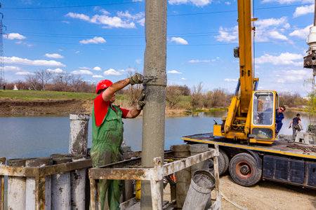 Rigger, worker with safety helmet disassembles pump canal for vertical charging, pouring fresh concrete into bridge foundation on river coast.の写真素材