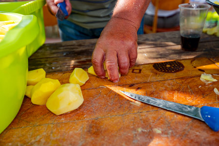 An elderly farmer cooker cuts potatoes using knife into slices on the wooden board, prepares it for cooking at the outdoor. Bowls are full of cut vegetableの写真素材