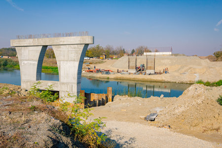 Large modern gray concrete bridge pillar in foundation surrounded by metal piles. Reinforced pole under construction is across river. Team of people worksの写真素材