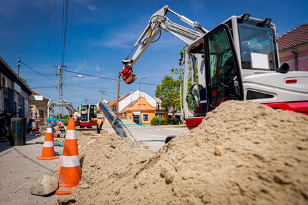 Several orange traffic cones with white fluorescent stripes are pointing for caution next to excavator as using straps to holding plumbing, plastic HDPE pipe of underground pipeline for water supply.の写真素材