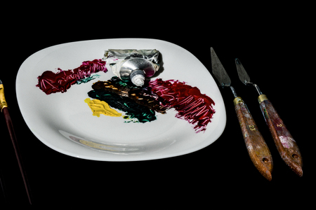 On a black background is a white porcelain plate. On the plate is a tube of paint and made color strokes. To the right of the plate are two spatulas for the artist, to the left are brushes.の写真素材