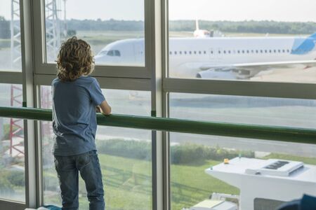 A blond boy with curly hair stands at a window in the airport building. Outside the window we could see the silhouette of the aircraft. Selective focus. Copy space.の写真素材