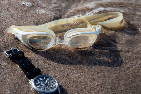Swimming goggles and a waterproof watch lie on the sand by the sea's shoreline. The sea wave fits the glasses. Copy space. Selective focus. Blurred motion. Defocus image. Blurred.の写真素材