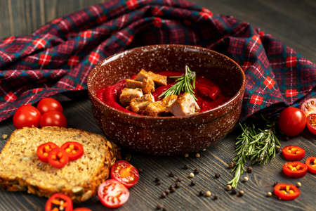 Thick tomato soup with meat slices in a ceramic plate. Next to the plate are a checkered serving napkin, bread, tomatoes, and a sprig of rosemary.の写真素材