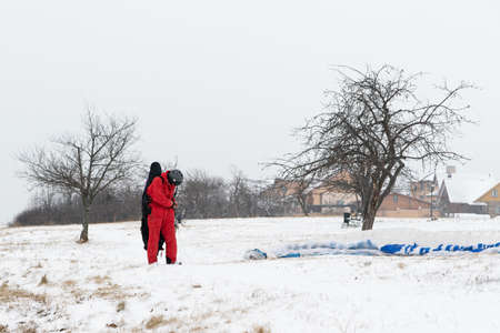 Winter. A pilot in a warm red jumpsuit is preparing for a paragliding flight. The wing is lying on...
