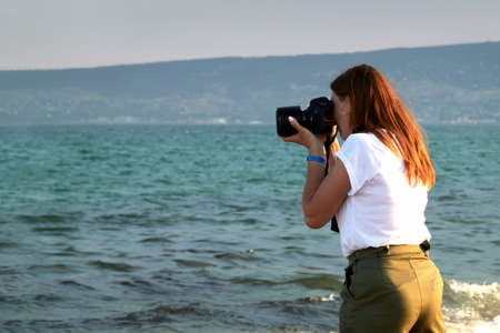 Crimea, Beregovoe, August 12, 2020: a young woman takes photos of a seascape with a camera. Selective focus.のeditorial素材