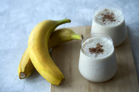 Two glasses with a milky banana cocktail on a bluish gray background. Next to the glasses are two...