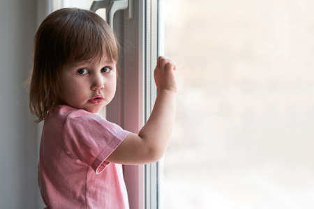 A little girl in home clothes at the window. Children's portrait.