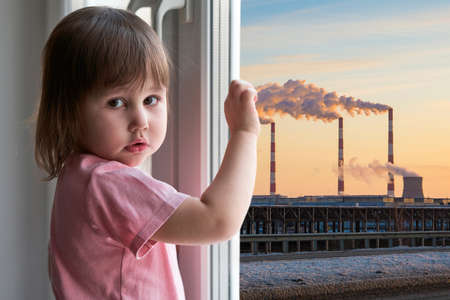A little girl with a sad face stands at the window, outside the window is a bleak technogenic urban landscape with smoking factory chimneys.の写真素材