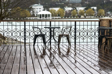 Summer cafe on the lake with mountain views. Behind the wrought-iron fence on the wooden deck is a table with two chairs it is raining. Copy space.の写真素材