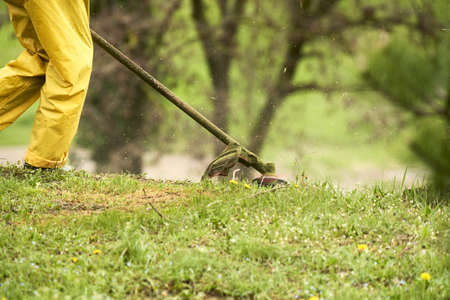 A fragment of a worker in a yellow uniform, lying in wait for a grass trimmer lawn. Grass dust flies...