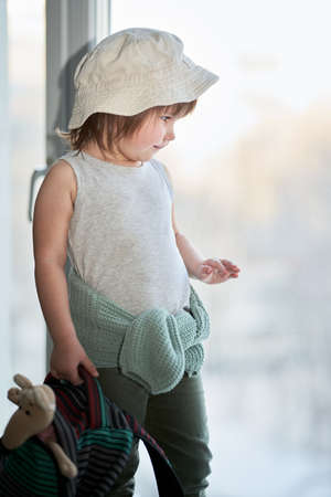 A little girl in a Panama hat with a backpack in her hand stands at the window. She was going to travel.の写真素材