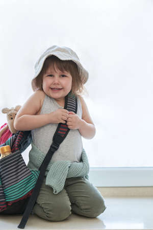 The little girl is emotionally happy to hear about the upcoming trip. She is sitting by the window with a backpack in which she has packed her favorite toys. Selective focus.の写真素材