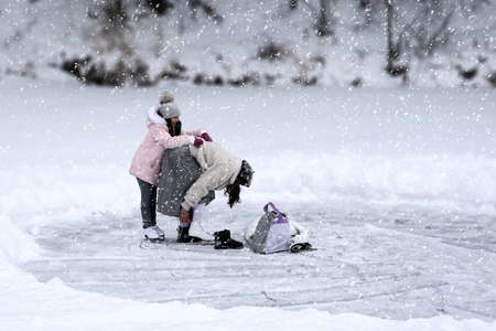 Russia, Naberezhnye Chelny, December 27, 2020: mother and daughter at the ice rink. A woman puts on skates, a girl on skates holds on to her mother. Snow is falling.のeditorial素材