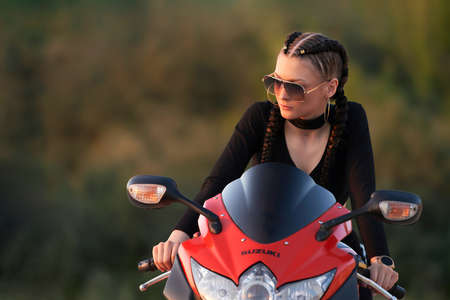 Russia, Naberezhnye Chelny, June 20, 2021: a girl wearing sunglasses on a red motorcycle. The girl's hair is braided in pigtails. The green background of the image is blurred.のeditorial素材