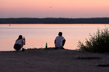 A guy and a girl are sitting on the shore of a lake at sunset. There is a bottle of beer next to the...