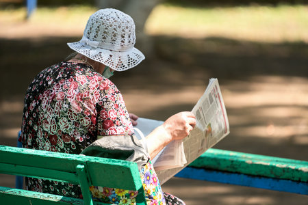 An elderly woman in a flirty knitted hat is sitting on a bench in the square and reading a newspaper. She has a medical mask on her face.の写真素材