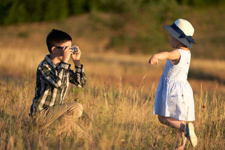 The older brother takes pictures of his sister with a camera in nature. A little girl in a straw hat willingly poses for a young photographer.の写真素材