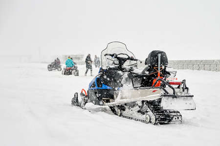 Winter entertainment. In the foreground is a snow-covered snowmobile without a driver, in the background people are riding quad bikes. Snow is falling. Selective focus.の写真素材