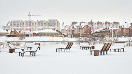Winter city landscape. Snow-covered benches on the river bank against the background of low-rise buildings and multi-storey construction.の写真素材