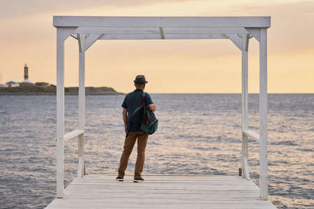 An adult man in a hat standing on a sea pier and looking straight. He admires the sea in the rays of the setting sun. Copy space.の写真素材