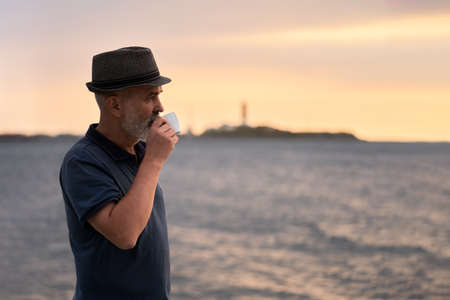 An adult gray-haired man in a hat on the beach with a cup of coffee. He looks at the sea in the rays...