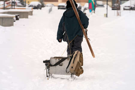 A tired fisherman returns from ice fishing. He pulls a sled with fishing gear. He has one ski on his back. Selective focus.の写真素材