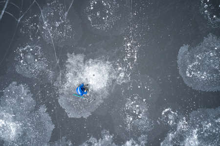 A lone fisherman is fishing on the thin, fragile ice of a reservoir covered with ice flowers. There is an ice drill nearby. Shooting from a drone.の写真素材