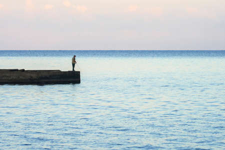 Seascape with a lonely fisherman fishing from the pier in the soft light of the setting sun. Copy space.の写真素材