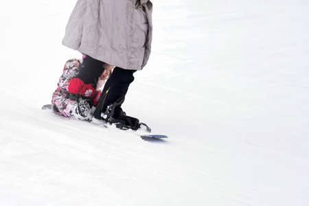 Russia, Fedotovo, February 28, 2021: A young woman goes down with a child on a snowboard from a mountain. The girl sitting on the board is holding on to her mother's leg. She's thrilled.のeditorial素材