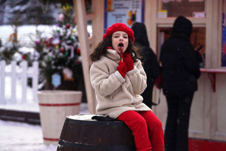 Russia, Moscow, December 20, 2021: Holiday Fair. A girl frozen in the cold licks a red lollipop on a stick. Selective focus.のeditorial素材