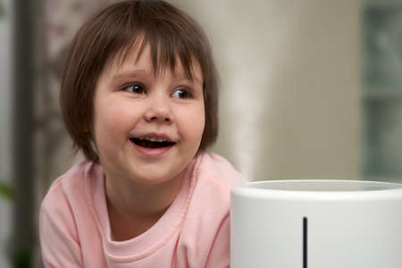 The little girl smiles happily, watching the jet of cold steam coming out of the humidifier. close-up.の写真素材