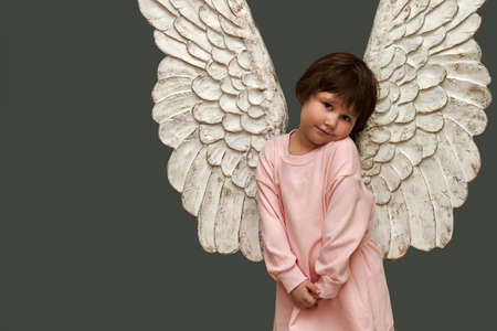 An embarrassed angel on a gray asparagus background. Cute little girl in pink dress with angel wings. copyspace.の写真素材